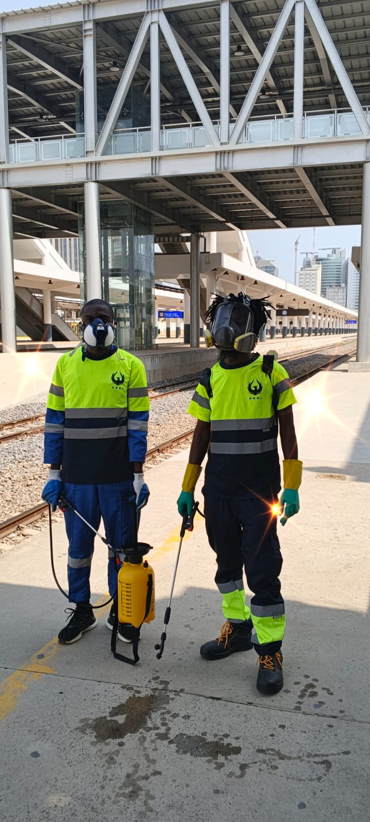 Dois técnicos da AKDL com fardamento refletor e pulverizador numa estação em Luanda